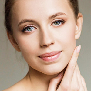 Close-up portrait of a young woman with clear skin and blue eyes gently touching her jawline.