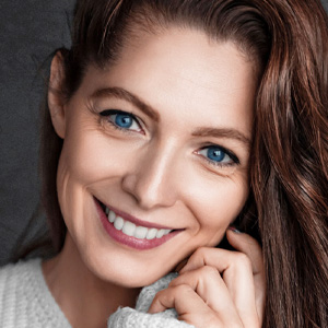Close-up portrait of a smiling woman with bright blue eyes and long brown hair wearing a white sweater against a dark background.
