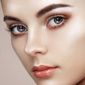 Close-up portrait of a woman with striking grey eyes, sculpted brows, and glossy peach lips showcasing luminous skin makeup.