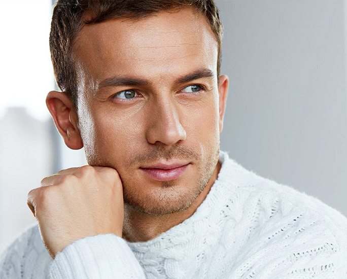 Close-up portrait of a thoughtful man with light stubble wearing a white cable-knit sweater looking off to the side.