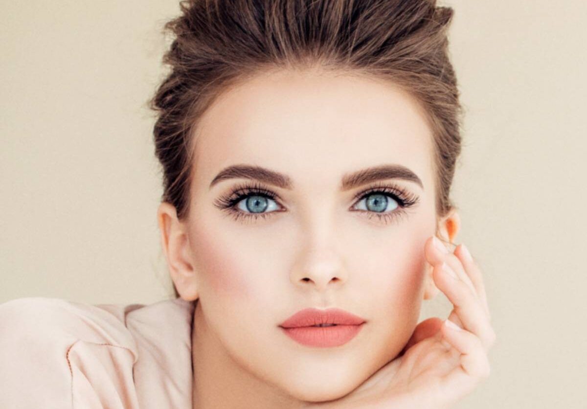 A woman with elegant makeup and bright blue eyes gazes intently, her chin resting on her hand. Her expression is calm and poised, exuding sophistication.